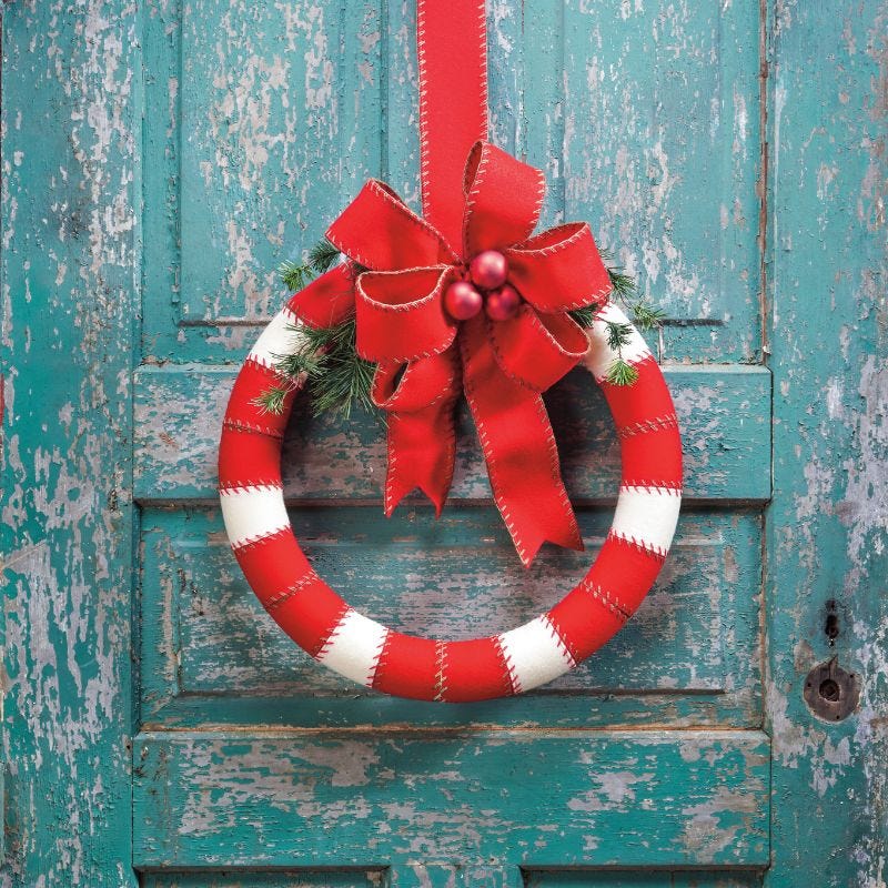 Foam wreath decorated with white and red ribbon, greenery, and a red bow with jingle bells displayed on a weathered door