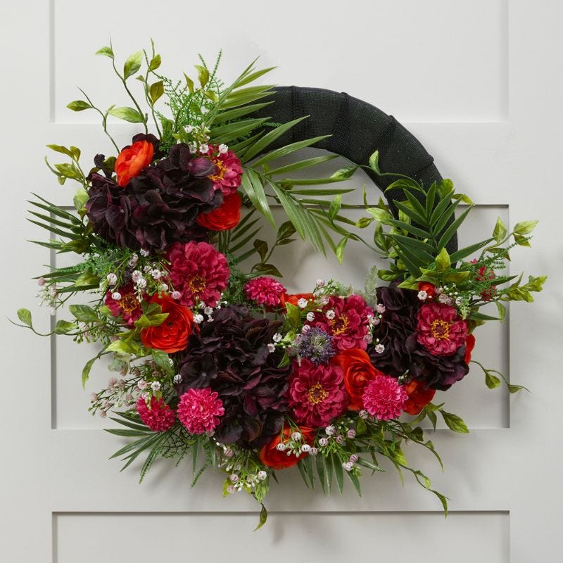 Red and dark purple flowers are arranged with greenery on a ribbon wrapped wreath form that is hung on a white door
